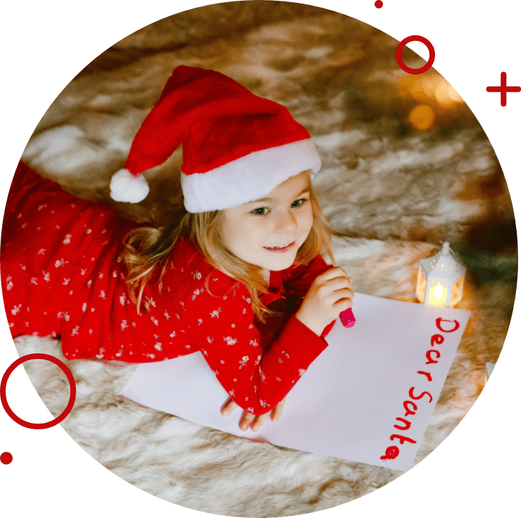Young girl wearing a Santa hat and red pajamas writing a 'Dear Santa' letter while lying on a cozy blanket with a small lantern nearby.
