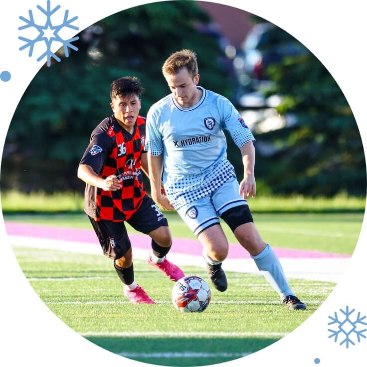 A soccer match with a player in a light blue Minnesota Blizzard FC jersey dribbling the ball, closely followed by a player in a red and black checkered jersey, on a green field under sunny conditions.